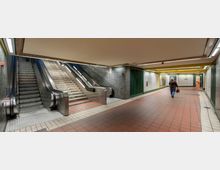 U-Bahn-Station mit einem breiten, gefliesten Gang, einer Rolltreppe und einer danebenliegenden Treppe aus Stein. Die Wände sind mit weißen Fliesen verkleidet, ergänzt durch grüne und gelbe architektonische Details.