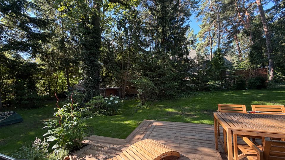 Holzterrasse mit Tisch, Stühlen und geschwungener Liege vor einem großen, gepflegten Garten mit Rasenfläche. Im Hintergrund stehen hohe Laub- und Nadelbäume sowie dichtes Strauchwerk, teilweise mit Blick auf ein Hausdach durch die Bäume.