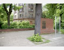 Ein historisches, mehrstöckiges Backsteingebäude mit Rundbögenfenstern, umgeben von Bäumen und dichter Vegetation. Im Vordergrund befindet sich eine Gedenkwand aus roten Ziegeln mit einer Metalltafel sowie eine transparente Glasstele, die auf einem gepflasterten Weg stehen.