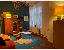 Wohnzimmer mit Holzboden, einer grünen Wand mit einem hölzernen Retro-Schrank und verschiedenen Kunstwerken sowie einem großen Fenster mit weißen Vorhängen. Im Raum stehen ein kleiner Couchtisch, eine orangefarbene Couch und mehrere Steh- und Tischlampen, die warmes Licht spenden.