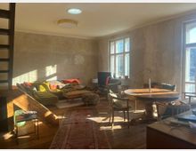 Ein gemütliches Wohnzimmer mit sichtbaren, unverputzten Wänden und Holzboden. Es verfügt über ein großes Sofa mit bunten Kissen, einen runden Holztisch mit Stühlen, mehrere Fenster, die viel Tageslicht hereinlassen, sowie eine Holztreppe im Vordergrund.