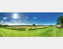 Weitläufige, gepflegte Rennbahn mit grünen Wiesen, Hecken und einzelnen Bäumen unter klarem, sonnigem Himmel. Im Hintergrund dichter Wald und am rechten Bildrand Gebäude mit rotem Dach und gepflastertem Weg.