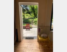 Blick aus einem Innenraum durch eine offene Tür auf eine geflieste Terrasse mit rotem Stuhl; dahinter führt ein schmaler Pfad mit Stufen in einen grünen Garten mit Bäumen und Zaun. Im Vordergrund sind Holzboden und rechts ein kleiner gelber Beistelltisch mit Radio zu sehen.