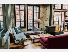 Gemütlicher Wohnbereich mit einem gemusterten Ecksofa, einem roten Sessel und einem Holztisch auf Holzboden, umgeben von Wänden aus Backstein. Große Fenster mit schwarzen Rahmen und ein Holzofen verleihen dem Raum ein industrielles und rustikales Ambiente.