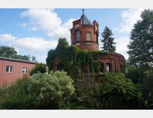 Historisches Backsteingebäude mit einem runden Turm, überwuchert von dichtem Efeu, umgeben von Bäumen und Sträuchern; daneben steht ein modernes, schlichtes Backsteingebäude.