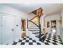 Heller Eingangsbereich mit schwarz-weiß kariertem Boden, einer geschwungenen schwarzen Holztreppe mit weißen Geländern und mehreren Türen zu angrenzenden Räumen. Dekorative Elemente wie ein goldener, giraffenförmiger Kleiderständer und Wandleuchter prägen das Ambiente.