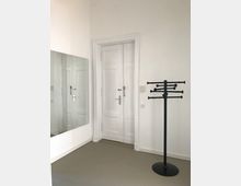 Minimalistischer Innenraum mit weißen Wänden und einer doppelten, weißen Tür mit klassischer Holzverkleidung. Links hängt ein großer rechteckiger Wandspiegel, rechts steht ein schwarzer Garderobenständer auf einem hellgrauen Boden.