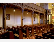 Innenansicht einer historischen Kirche mit Holzbänken und verzierten Säulen. Beleuchtung durch hängende Lampen, Wandkerzenhalter und kleine Tafeln mit Liednummern an den Wänden, helle Wände und eine Empore im oberen Bereich.