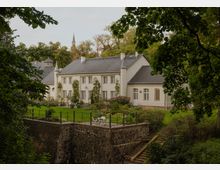 Blick auf ein helles, schlossähnliches Gebäude mit grauem Satteldach, vielen Sprossenfenstern und kleinen Türmchen, umgeben von Bäumen. Davor liegt ein gepflegter Garten mit Sitzgelegenheiten auf einer erhöhten Terrasse über einer Natursteinmauer.