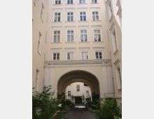 Mehrstöckiges Wohngebäude mit hellbeiger Fassade, weiß gerahmten Fenstern und einem großen Rundbogen, der einen Durchgang zu einem Innenhof mit Pflanzen und einem weiteren Gebäude bildet. Ein lateinischer Schriftzug mit Jahreszahlen ist über dem Bogen angebracht.