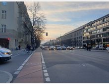 Straßenszene bei Sonnenuntergang mit mehrspuriger Straße in einer Stadt. Links und rechts säumen mehrstöckige Gebäude mit schlichten Fassaden die Straße, während Autos, Fahrräder und Fußgänger den Verkehr teilen.