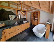 Modernes Badezimmer mit freistehender weißer Badewanne, dunklem Schieferboden und Holzinterieur. Eine große Holz-Waschtischanlage mit integriertem Spiegel und Waschbecken sowie offene Holzregale prägen den Raum, der durch sichtbare Dachbalken und ein geneigtes Holzgebälk geprägt ist.