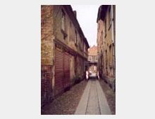 Schmaler gepflasterter Durchgang zwischen zwei alten, verwitterten Gebäudezeilen mit Backstein- und Putzfassaden; im Hintergrund ein Bogen mit Blick auf ein weiteres Gebäude.