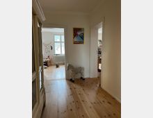 Flur in einer Wohnung mit hellem Holzboden, weißen Wänden und zwei offenen Türen, die in angrenzende Räume führen. An der Wand hängt ein Poster, und auf dem Boden steht ein gepunktetes Spielzeugpferd.