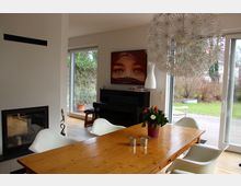 Esszimmer mit einem großen Holztisch und weißen Stühlen, daneben ein Kamin in einer weißen Wand. Im Hintergrund ein schwarzes Klavier vor einer Wand mit Kunstwerk, sowie große Fenster und eine Schiebetür mit Blick auf den Garten.