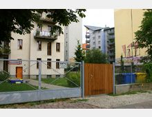 Mehrstöckige Wohnhäuser mit Balkonen und großen Fenstern, im Vordergrund ein Zaun aus Drahtgeflecht mit Holztor und Hinweisschildern, dahinter ein kleiner Rasenbereich und bunte Mülltonnen. Eine gelbe Hauswand zeigt Graffiti-Kunst.