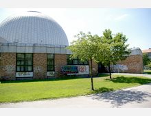 Gebäude mit einer großen, weißen Kuppel und einer Fassade aus Backstein mit mehreren Fenstern, die teilweise von Graffiti bedeckt sind. Im Vordergrund befindet sich eine Rasenfläche mit zwei kleinen Bäumen und einem gepflasterten Weg.