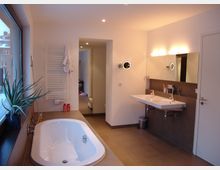 Helles, modernes Badezimmer mit einer weißen Badewanne, einem großen Fenster links, einem Doppelwaschbecken mit Spiegel rechts und einem Handtuchheizkörper an der Wand. Die Bodenfliesen und Wände sind in neutralen Farbtönen gehalten, und eine Aloe-Pflanze steht auf der Fensterbank.