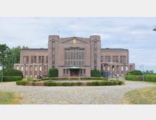 Backstein-Gebäude im monumentalen Stil mit symmetrischer Fassade, großen Fensterflächen und einem hervorgehobenen Eingangsbereich mit Vordach. Der Platz davor ist mit Kopfsteinpflaster gestaltet, umgeben von gepflegten Hecken und Lampen.