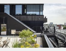 Moderne Dachterrasse mit Sitzbereich, Pflanzenbeeten und Treppe, die zu einem Gebäude mit dunkler Holz- und Glasfassade führt; im Hintergrund städtische Umgebung mit mehreren Gebäuden.