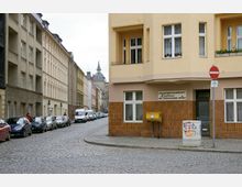 Straßenszene mit gepflasterter Straße und mehrstöckigen Wohngebäuden im Altbaustil. Ein gelbes Eckgebäude mit einem Briefkasten und einem Schild für Krankentransport-Dienste steht im Vordergrund, während Autos dicht am Straßenrand geparkt sind.