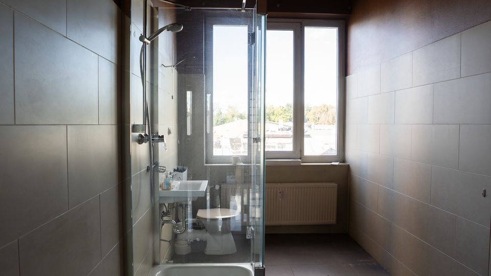 Schmales, modern gefliestes Badezimmer mit Badewanne und Glas-Duschabtrennung sowie kleinem Waschbecken und Toilette. Am Ende des Raums befindet sich ein großes Fenster mit Heizkörper darunter, das viel Tageslicht hereinlässt.