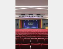 Großer Veranstaltungsraum mit mehreren Reihen roter Sitze, ausgerichtet auf eine Bühne mit geschlossenem Vorhang in Blau und Rot; über der Bühne ist Schriftzug "Urania Berlin" sichtbar, und die Decke ist mit modernen, linearen Beleuchtungselementen gestaltet.