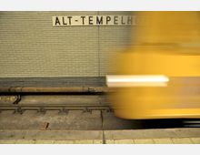 U-Bahnhof mit gelber Ziegelwand und der Aufschrift „Alt-Tempelhof“, im Vordergrund verschwommen ein vorbeifahrender gelber U-Bahn-Wagen auf den Gleisen.