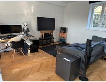 Wohnzimmer mit modernem Arbeitsbereich und Unterhaltungsecke. Ein Schreibtisch mit Computer, Monitor und Dekorfigur steht neben einem großen Fernseher auf einem rustikalen TV-Regal; im Vordergrund ein graues Sofa auf Parkettboden vor einem schwarzen Teppich, mit Tageslicht durch ein seitliches Fenster.