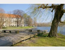 Uferbereich mit hölzernem Steg und Sitzmöglichkeiten, daneben ein großer Baum mit überhängenden Zweigen. Im Hintergrund ein Gebäude mit rotem Ziegeldach sowie ein Fluss oder See, umgeben von kahlen Bäumen und vereinzelt modernen Gebäuden.