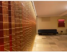 Moderner Innenbereich mit einer dekorativen, goldfarbenen Metallwand auf der linken Seite, die aus vertikalen und wellenförmigen Elementen besteht. Rechts befindet sich ein schwarzes Ledersofa, ein Mülleimer und ein rot hinterlegter Spiegel an der hellen Wand, der Boden ist mit großformatigen beige-gemusterten Fliesen ausgelegt.