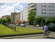Straßenszene in einer städtischen Umgebung mit mehrstöckigen Wohngebäuden, einem Fußweg, geparkten Autos und kleinen Geschäften mit Markisen. Im Vordergrund eine grasbewachsene Fläche mit Zaun, im Hintergrund Bäume und Verkehr auf der Straße.