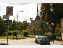 Straßenszene mit parkenden Autos und einem Wegweisschild, im Hintergrund eine Kirche mit spitzem Turm und gotischen Fenstern, umgeben von Bäumen und Grünflächen.