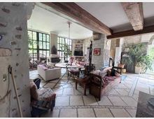Großzügiger Wohnbereich im rustikalen Stil mit sichtbaren Holzbalken, Steinwänden und hellem Natursteinboden in geometrischem Muster. Große Fensterfronten, ein Kamin und mehrere Sofas und Sitzgruppen prägen den offenen Raum mit Blick ins Grüne.