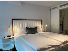 Modernes Hotelzimmer mit einem Doppelbett, das ein weiß-gepolstertes Kopfteil mit Holzrahmen hat. Neben dem Bett stehen zwei Nachttische mit Lampen, und im Hintergrund ist ein offener Kleiderschrank zu sehen.