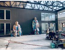 Eine helle, industriell gestaltete Halle mit einem transparenten Dach und Metallträgern. Im Vordergrund stehen zwei bunte, großformatige Skulpturen von Figuren vor einer grauen Wand mit Fenstern und einer Tür; rechts ist ein Arbeitsplatz mit Regalen, Flaschen und Utensilien zu sehen.