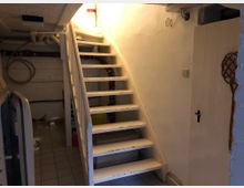 Treppenaufgang in einem Kellerraum mit weiß gestrichenen Wänden und Holztreppenstufen, einem Fliesenboden und sichtbaren Rohren an der Decke und Wand. Rechts befindet sich eine weiße Tür mit einem geflochtenen Deko-Element.