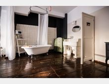 Helles Badezimmer mit freistehender weißer Badewanne auf glänzendem Holzboden, umgeben von einem Duschvorhang. Im Hintergrund sind weiße Wandfliesen, ein Waschbecken mit ovalem Spiegel sowie eine grüne Trennwand mit Blumenmotiven zu sehen.