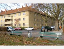 Wohngebäude in einem städtischen Umfeld mit gelb-beigem Putz und roten Ziegeldächern. Im Vordergrund sind eine Straße mit geparkten Autos, zwei grüne Bänke, ein Müllbehälter und vereinzelt Laubbäume zu sehen.