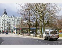 Straßenszene mit dem U-Bahnhof Südstern im Vordergrund und einem weißen Altbau mit Kuppelturm im Hintergrund. Im Bild sind außerdem Bäume ohne Laub, geparkte Fahrräder, ein graffitibemalter Lieferwagen und eine belebte Kreuzung zu sehen.