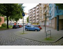 Straßenszene in einem urbanen Wohngebiet mit mehrstöckigen Wohnhäusern, einem kleinen Parkplatz mit parkenden Autos und einer gepflasterten Straße. Links stehen Bäume, rechts führt ein Gehweg an den Gebäuden entlang, und ein Straßenschild weist auf die Schillerpromenade hin.