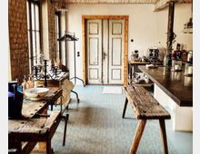 Rustikaler Innenraum mit einer offenen Küche, an einer Seite eine Arbeitsplatte aus Metall und Holz. Im Vordergrund ein Holztisch und eine Bank, im Hintergrund eine alte Doppeltür mit Holzrahmen sowie Backsteinwände und große Fenster.