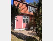Rotes Holzhaus mit Satteldach aus roten Ziegeln, einem verzierten Fenster mit Gitter und einer Holztür rechts. Der Bereich vor dem Haus ist kiesbedeckt, umrahmt von Bäumen und Sträuchern.