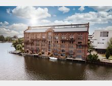 Backsteingebäude im industriellen Stil mit mehreren Etagen und Balkonen, direkt an einem Flussufer gelegen. Im Vordergrund befinden sich ein Steg und ein kleines Boot, während der Hintergrund von Bäumen und Nachbargebäuden geprägt ist.
