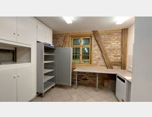 Kleine Küche mit Backsteinwand und sichtbaren Holzbalken, ausgestattet mit weißen Schränken, einem geöffneten Kühlschrank, einer Arbeitsplatte und einer Spülmaschine. Die Fensterrahmen aus Holz lassen Tageslicht einfallen.