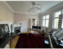 Wohnzimmer mit klassischem Kamin, einem Klavier, großem Teppich und Büromöbeln. Die Fenster sind hoch und lassen viel Tageslicht in den Raum, während im Hintergrund ein weißes Regal Bücher und Dekorationsgegenstände beherbergt.