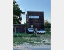 Ein modernes, zweistöckiges Haus mit einer Fassade aus dunklem Holz, großen Fenstern im oberen Stockwerk und flachem Dach. Davor stehen zwei geparkte Autos, ein weißer Geländewagen und ein weißes Cabrio, und das Grundstück ist von einem einfachen Holzzaun umgeben, mit Bäumen im Hintergrund.