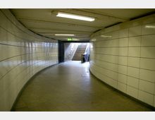 Ein U-Bahn-Tunnel mit geschwungenen, weiß gefliesten Wänden, beleuchtet durch rechteckige Deckenleuchten. Am Ende des Tunnels führen Treppen und eine Rolltreppe nach oben in einen beleuchteten Bereich.