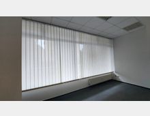 Ein leerer Büroraum mit einer großen Fensterfront, die mit vertikalen, weißen Lamellenjalousien bedeckt ist. Der Raum hat einen weiß gestrichenen Innenraum, eine abgehängte Decke mit Beleuchtungselementen und einen Heizkörper unter dem Fenster.