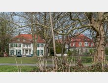 Zwei historische Häuser mit roten Ziegeldächern und weißen Fassaden, umgeben von einem Park mit großen, blattlosen Bäumen im Vordergrund. Ein Gehweg führt durch das parkähnliche Gelände, und vereinzelt sind Personen zu sehen.